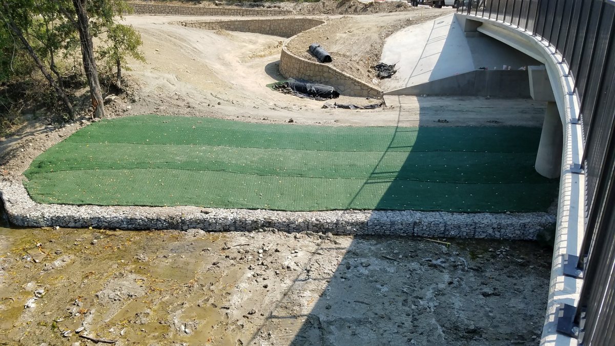 Green turf reinforcement mat anchored across a channel bank at a bridge outfall, edged with gabion stone